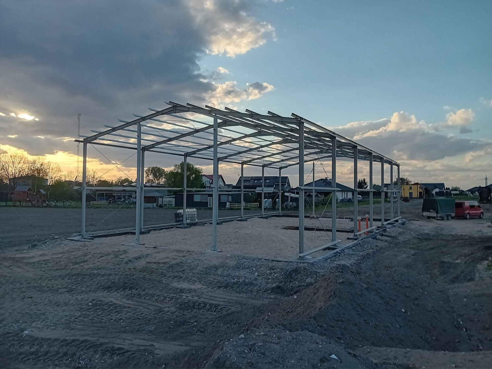 Konstrukcja stalowa hali w trakcie montażu, widok z zewnątrz na tle zachodzącego słońca i pochmurnego nieba. Widoczne elementy mocujące i teren budowy.