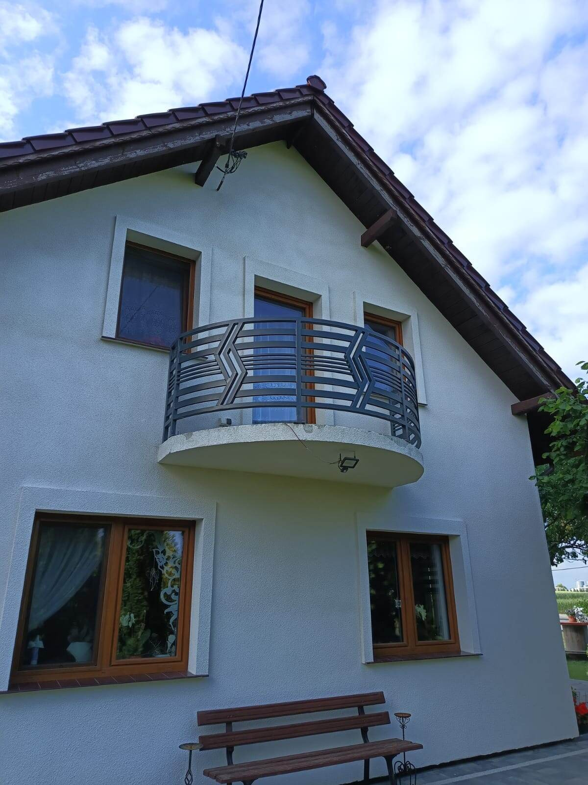 Biały dom z balkonem i nowoczesną, metalową balustradą o geometrycznym wzorze. Ujęcie z dołu, widoczny fragment dachu i niebieskie niebo z chmurami.