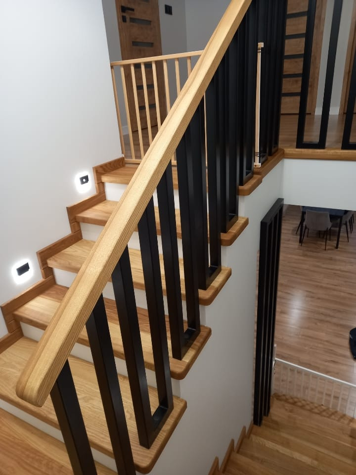 Nowoczesne schody z drewnianymi stopniami i czarną, metalową balustradą. Widoczne oświetlenie schodowe i fragment wnętrza domu z drewnianą podłogą.