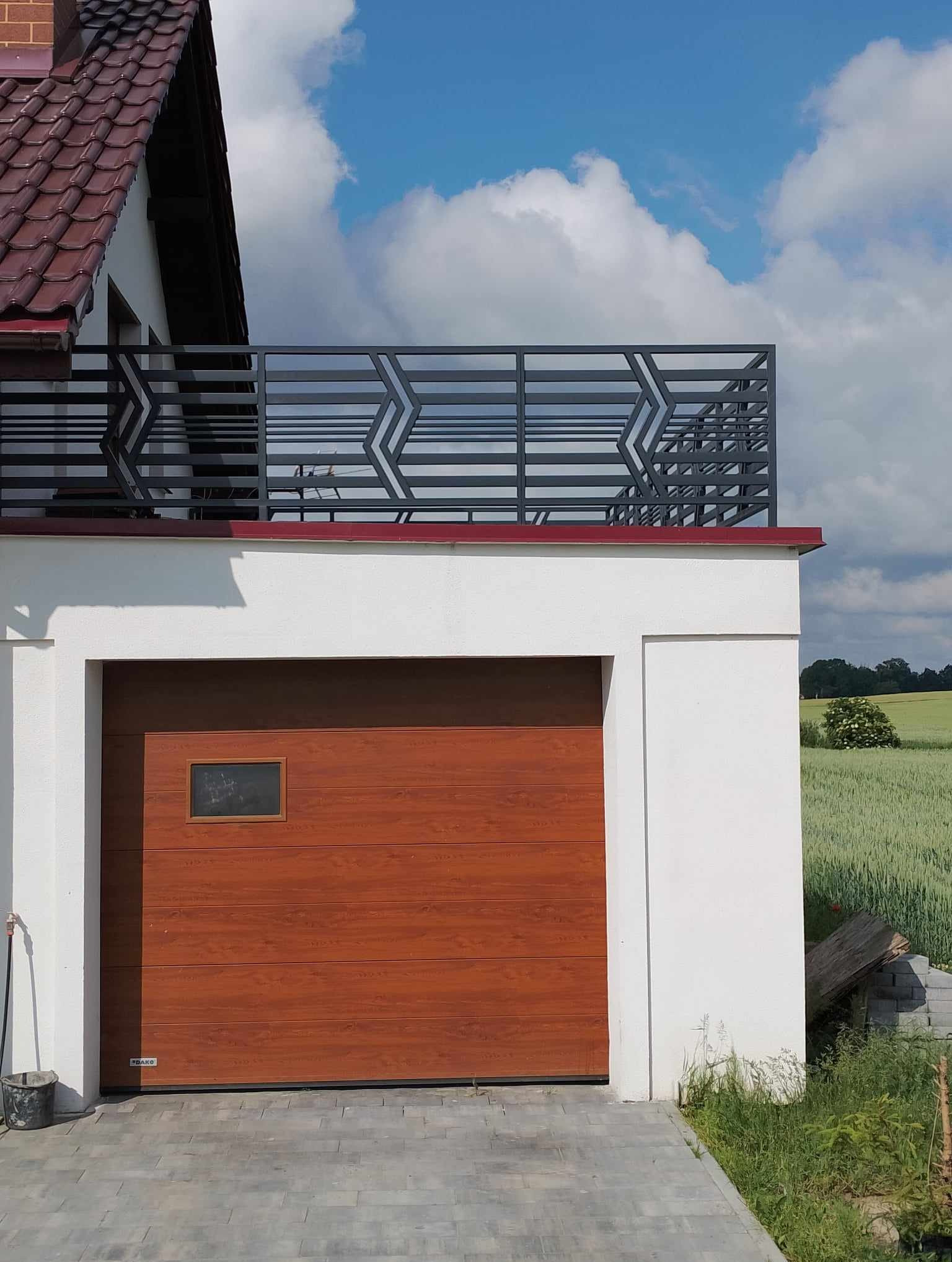 Nowoczesna brama garażowa z drewnianym wykończeniem i metalowa balustrada balkonowa z geometrycznym wzorem na tle błękitnego nieba i zielonego pola.