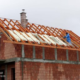 Darek Surgota - Prace izolacyjne na poddaszu domu w trakcie budowy. Widoczna biała pianka izolacyjna między krokwiami i pracownik w kombinezonie aplikujący ją. Ceglana konstrukcja budynku.