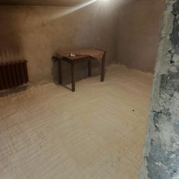 Darek Surgota - Pokój po aplikacji izolacji pianką PUR na podłodze, widoczny fragment ściany z surowej cegły, stary grzejnik i stół. Oświetlenie żarówką.
