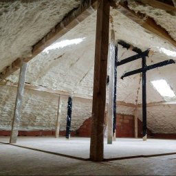 Darek Surgota - Poddasze z widoczną warstwą izolacji natryskowej, zabezpieczone elementy konstrukcyjne folią. Widoczne okno dachowe i fragment ściany z cegły.