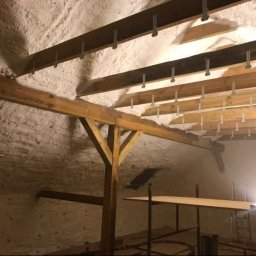 Darek Surgota - Poddasze z drewnianą konstrukcją i zaaplikowaną pianą izolacyjną, widoczne elementy mocujące. Oświetlenie podkreśla strukturę piany.