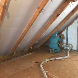 Darek Surgota - Pracownik w kombinezonie aplikuje pianę izolacyjną na poddaszu, widoczne drewniane krokwie i fragmenty ocieplonej ściany. Wąż doprowadzający pianę leży na podłodze.