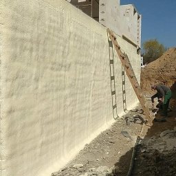 Darek Surgota - Izolacja fundamentów pianką: pracownik aplikuje pianę na ścianę fundamentu z użyciem drabiny. Widoczny wykop i fragment budynku w trakcie budowy.