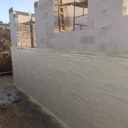 Darek Surgota - Ściana fundamentowa z bloczków, zabezpieczona warstwą izolacyjną w kolorze beżowym. Widoczne rusztowania i wykop ziemny. Budowa w trakcie realizacji.