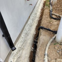 Darek Surgota - Izolacja fundamentów pianką PUR w wykopie przy ścianie budynku. Widoczne rury i instalacje w ziemi. Biała pianka szczelnie wypełnia przestrzeń.