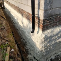 Darek Surgota - Izolacja fundamentu pianką PUR, widoczny róg budynku z nałożoną warstwą piany, obok wykop z instalacją. Detal: czarna rura spustowa przy ścianie.