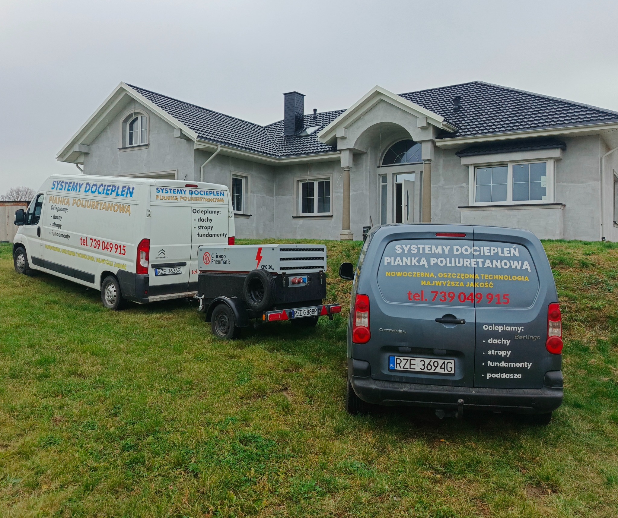 Dwa busy i przyczepa z logo firmy ocieplającej pianką PUR stoją na trawniku przed domem w trakcie budowy. Reklama usług na pojazdach: dachy, stropy, fundamenty, poddasza.