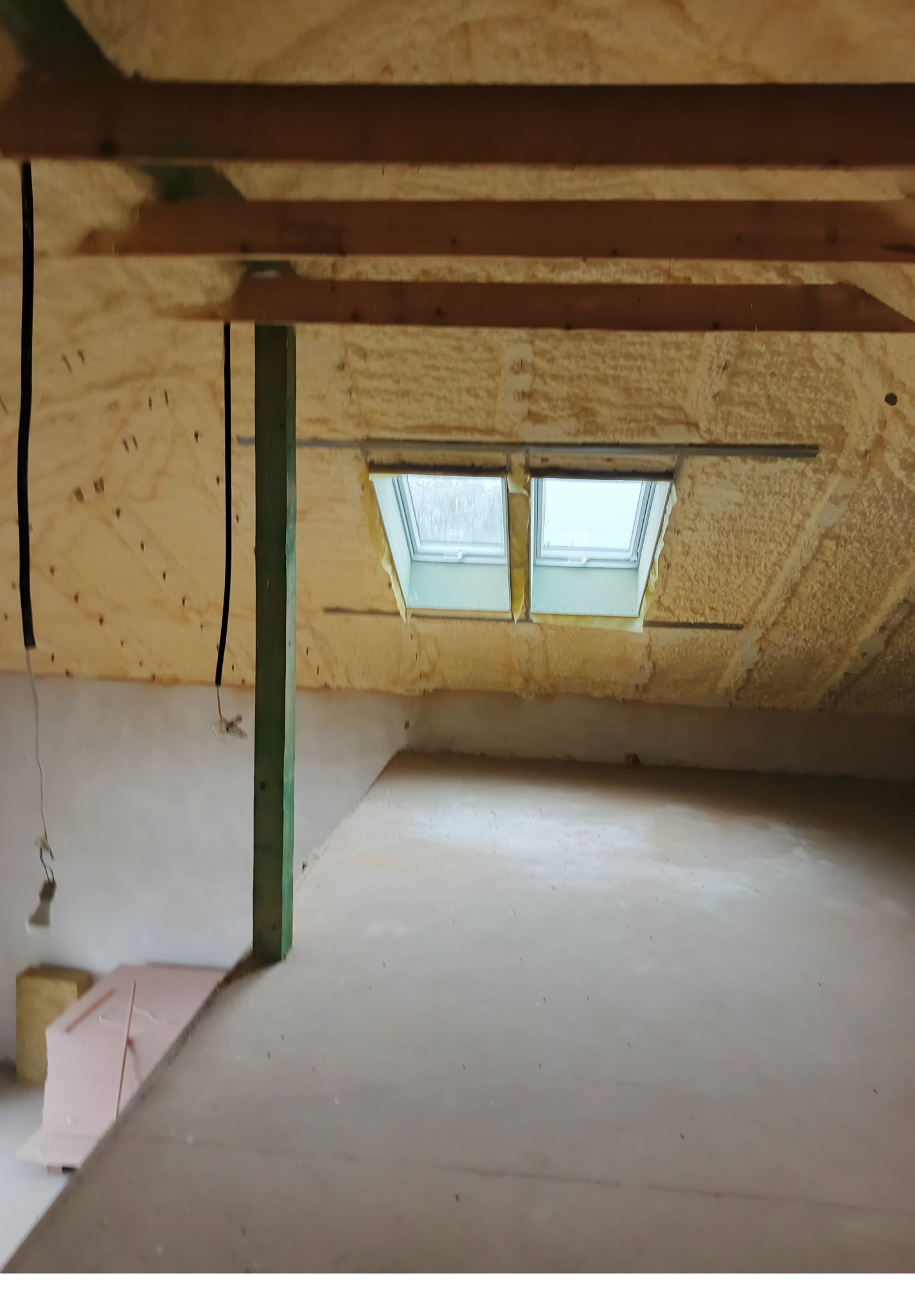 Poddasze z oknami dachowymi po ociepleniu pianką PUR, widoczne drewniane belki konstrukcyjne i fragment podłogi. Izolacja natryskowa o nieregularnej fakturze.