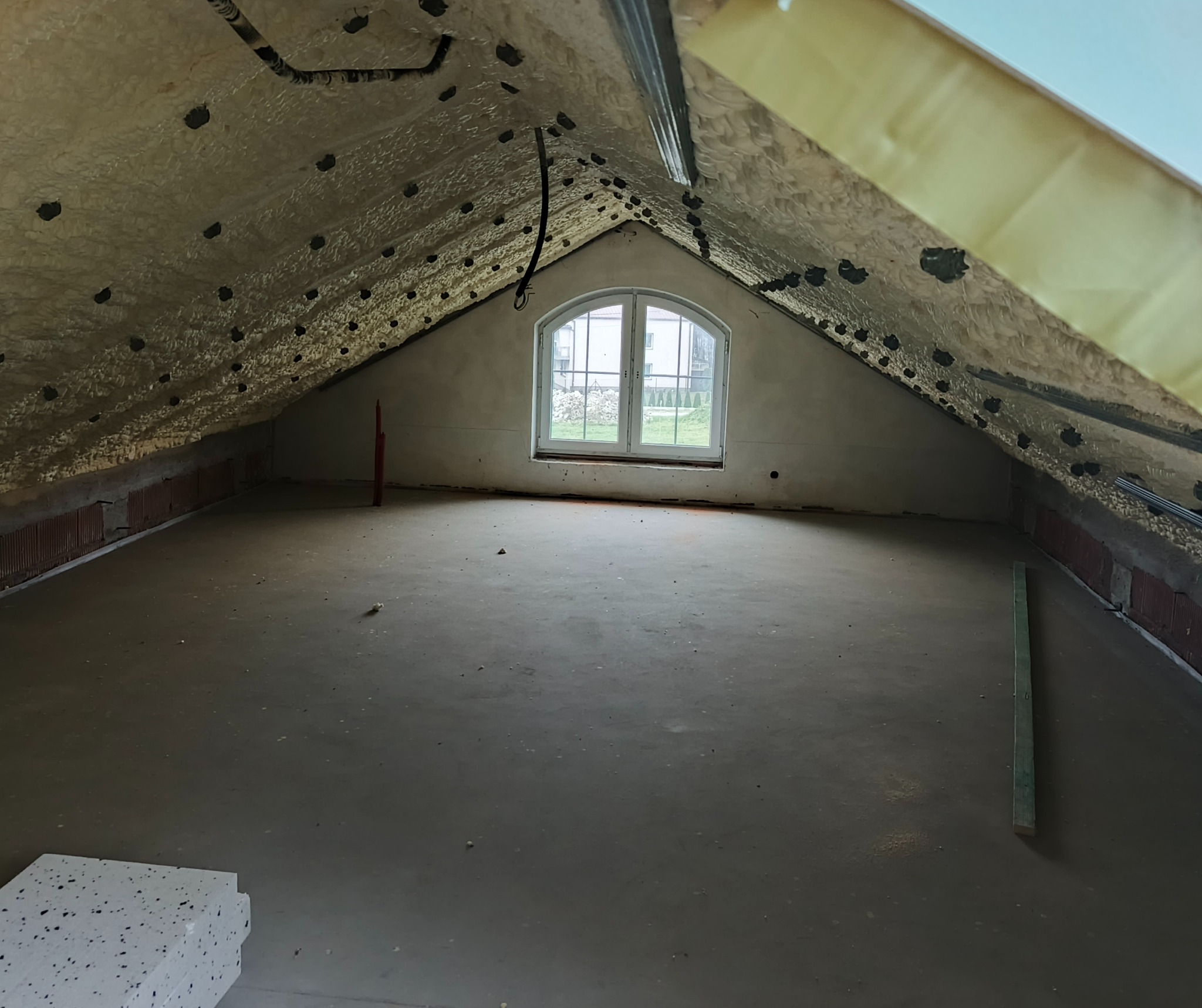 Poddasze po ociepleniu pianką PUR, widoczne czarne punkty po aplikacji. Okno w szczycie, surowa podłoga. Widok na zewnątrz przez okno.