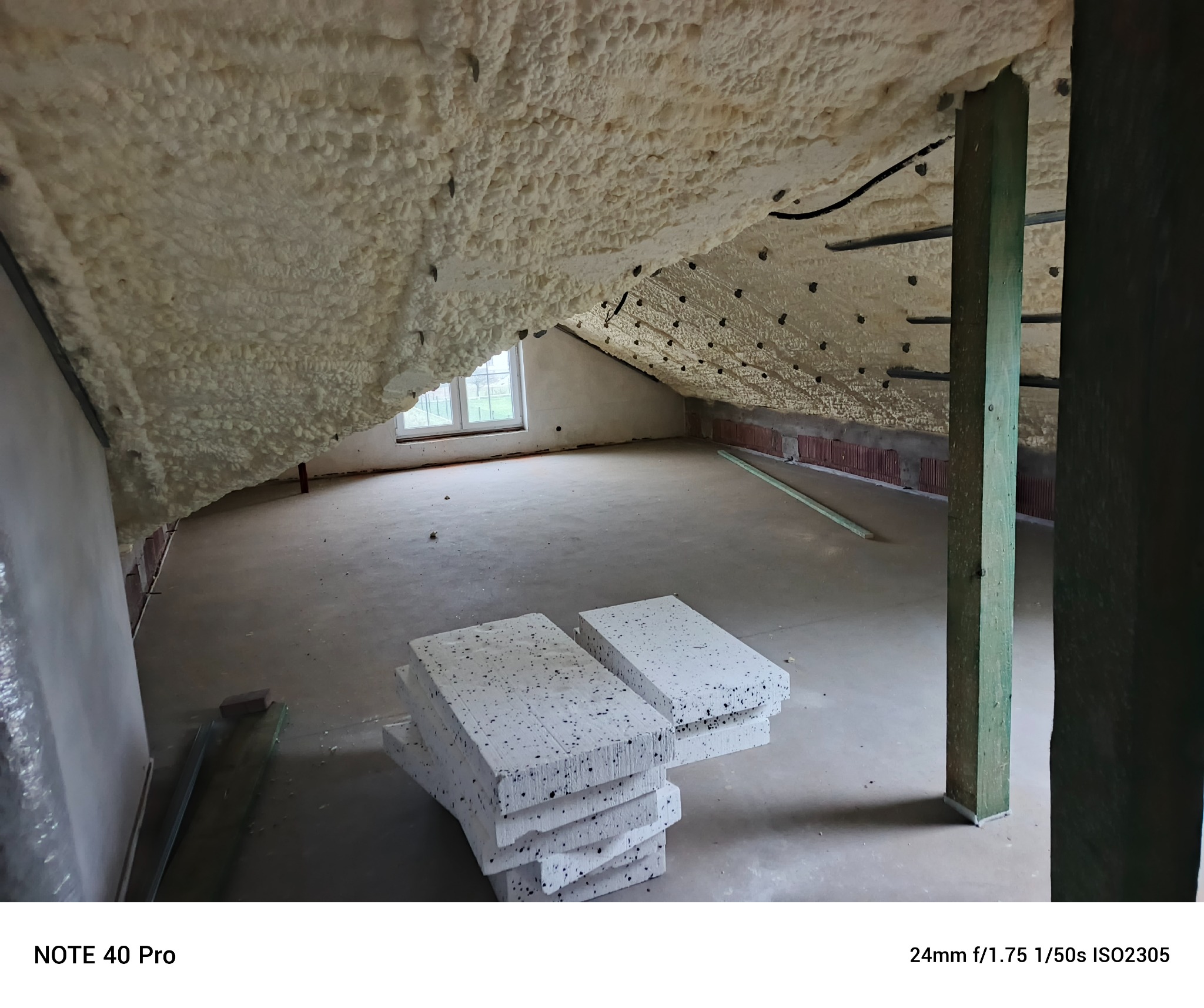 Poddasze po ociepleniu pianką PUR, widoczne krokwie i fragment okna. Na podłodze leżą płyty styropianowe. Ocieplenie wykonane natryskowo, tworzy jednolitą warstwę.