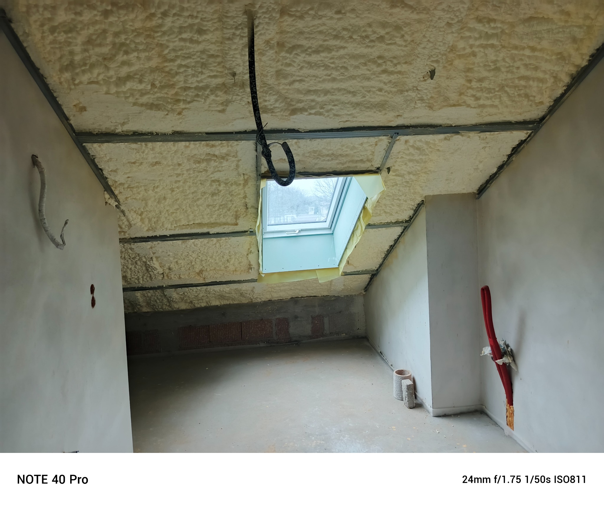 Poddasze po ociepleniu pianką PUR, widoczne metalowe profile i okno dachowe. Na ścianach instalacje elektryczne i hydrauliczne przed wykończeniem.