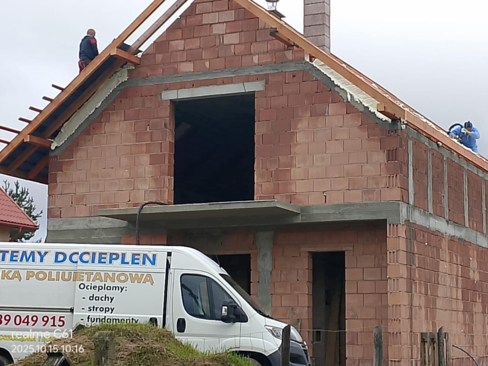 Budowa domu z czerwonej cegły, widoczni pracownicy na dachu podczas prac izolacyjnych. Na pierwszym planie samochód z reklamą ociepleń.