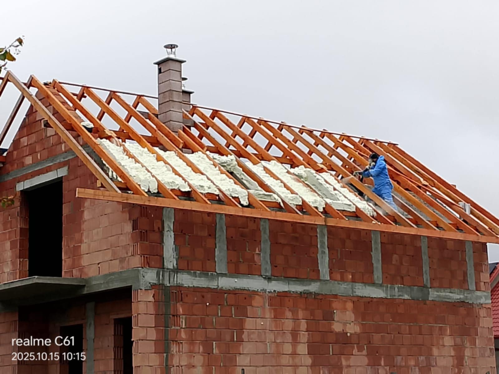 Prace izolacyjne na poddaszu domu w trakcie budowy. Widoczna biała pianka izolacyjna między krokwiami i pracownik w kombinezonie aplikujący ją. Ceglana konstrukcja budynku.