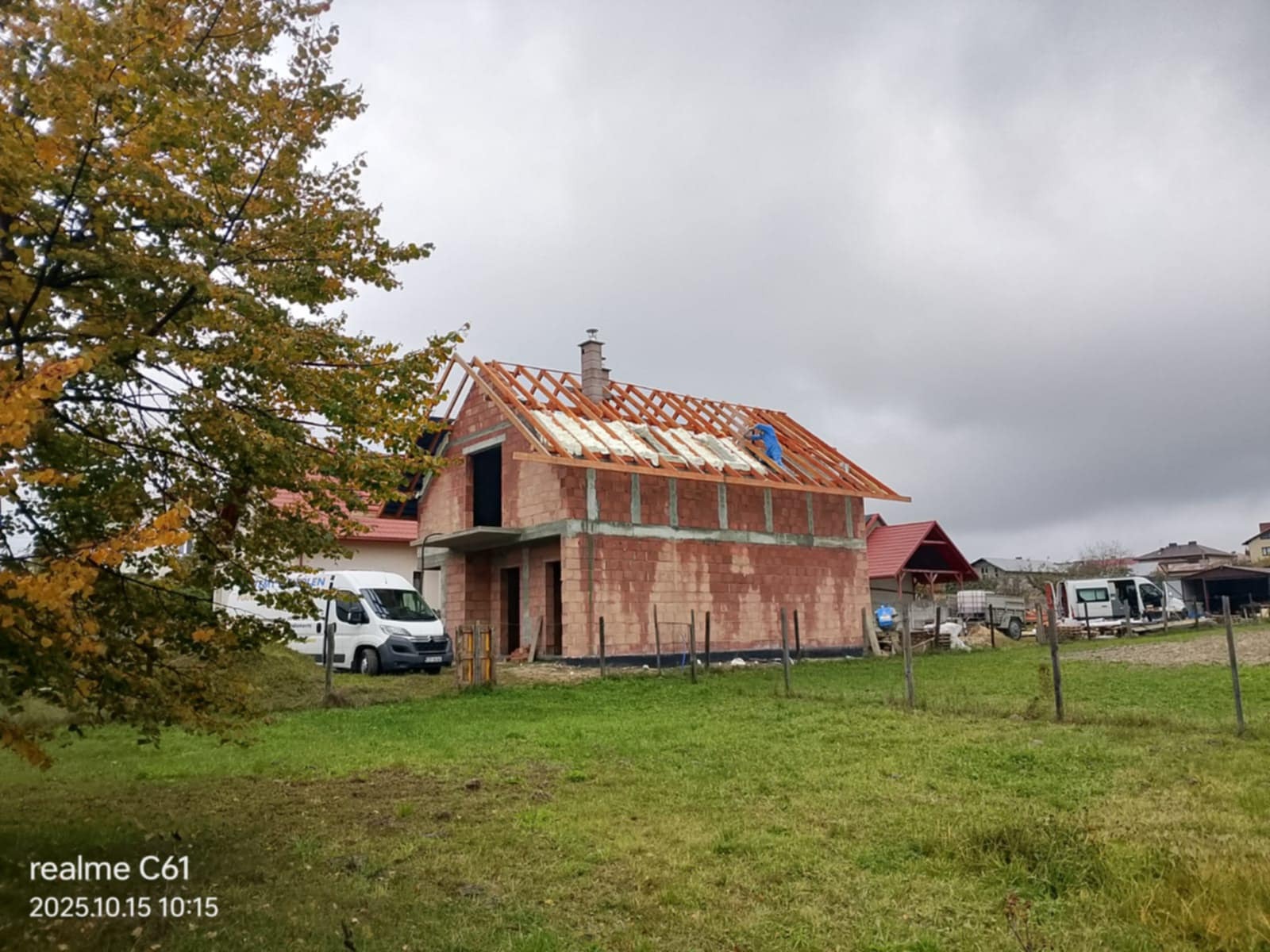 Budowa domu jednorodzinnego w trakcie ocieplania dachu. Widoczne krokwie, izolacja i pracownik w niebieskim kombinezonie. Na pierwszym planie trawa i drzewo.
