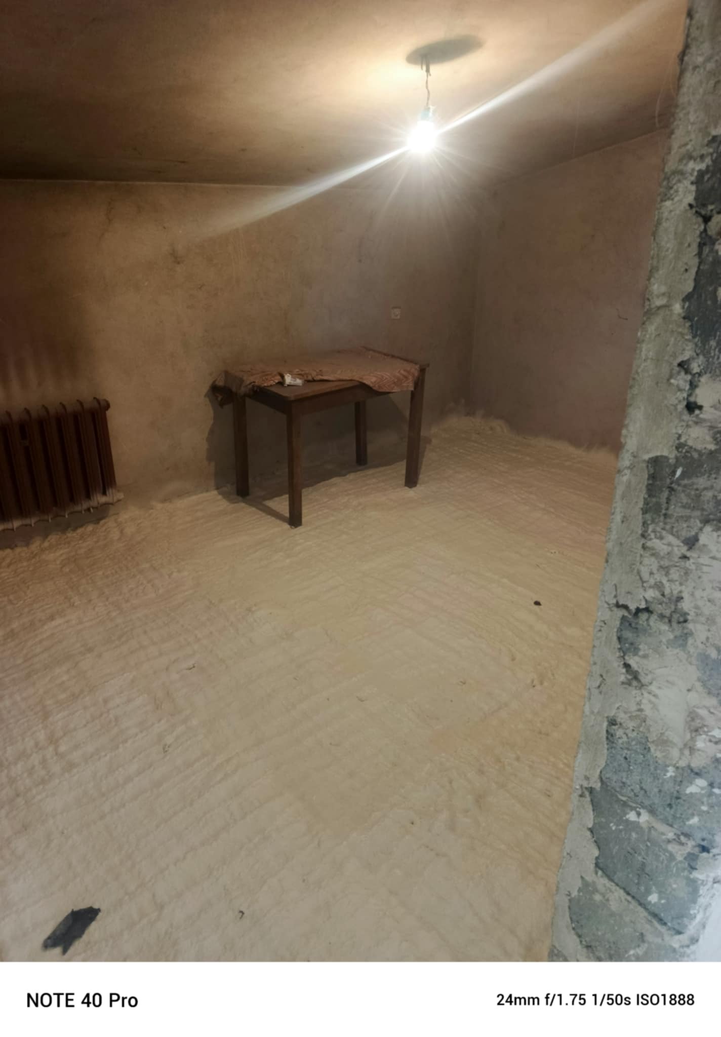 Pokój po aplikacji izolacji pianką PUR na podłodze, widoczny fragment ściany z surowej cegły, stary grzejnik i stół. Oświetlenie żarówką.