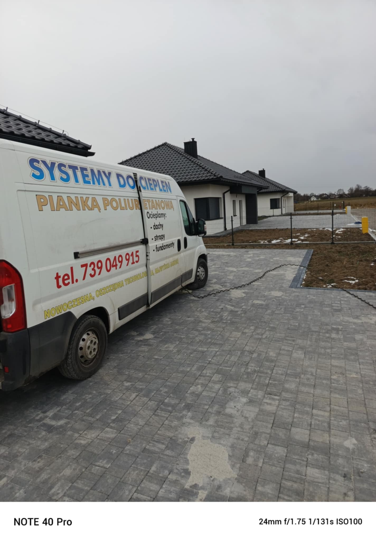 Biały bus z reklamą systemów dociepleń pianką poliuretanową na tle nowoczesnego domu jednorodzinnego z czarnym dachem i szarym brukowanym podjazdem.