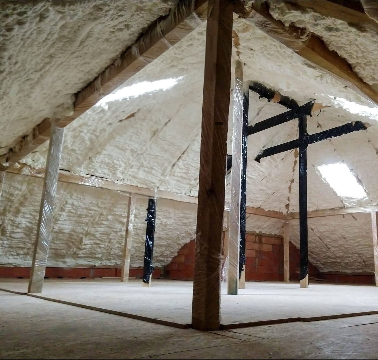 Poddasze z widoczną warstwą izolacji natryskowej, zabezpieczone elementy konstrukcyjne folią. Widoczne okno dachowe i fragment ściany z cegły.