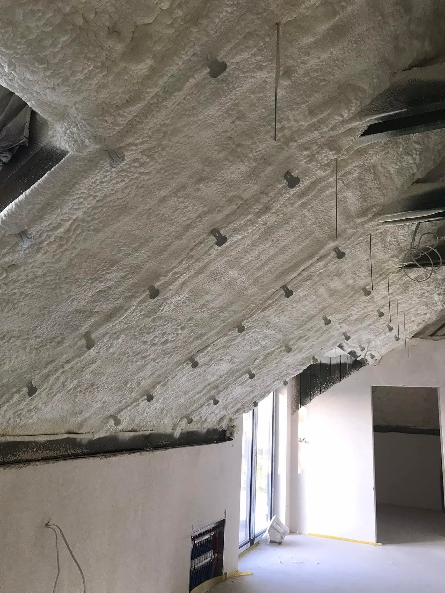 Poddasze po natrysku pianki PUR, widoczne metalowe elementy mocujące. Biała, nierówna powierzchnia izolacji pokrywa skośny dach. W tle okno i fragment ściany.