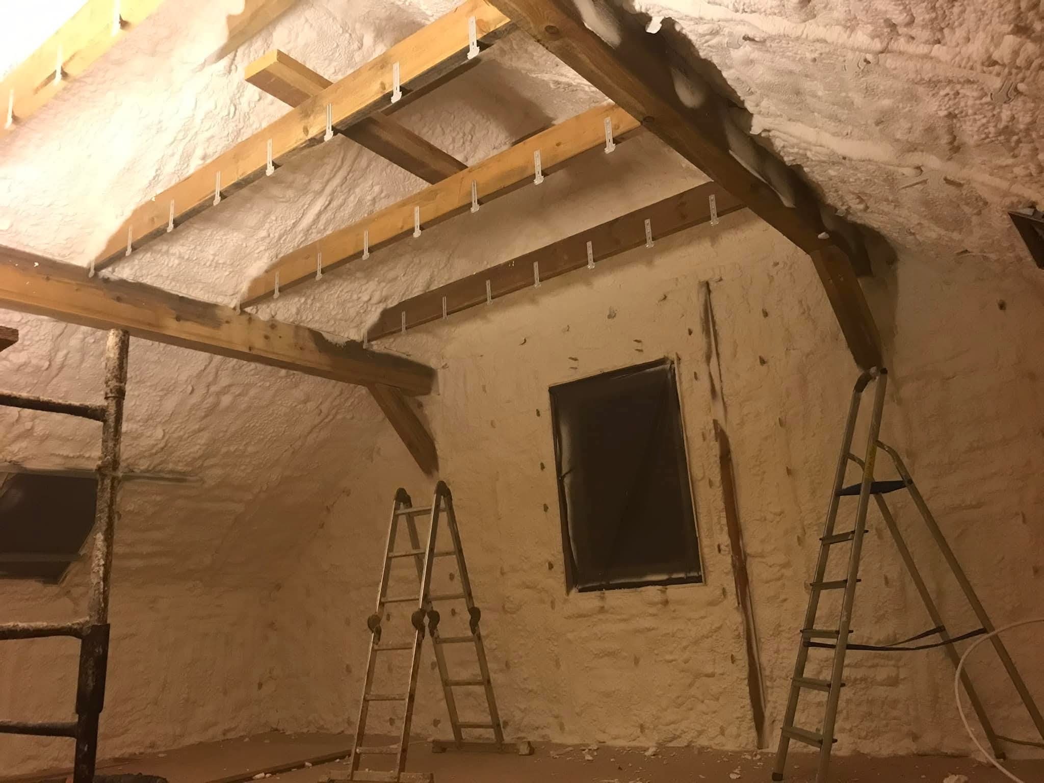 Poddasze po natrysku pianki PUR, widoczne drewniane elementy konstrukcyjne z plastikowymi uchwytami, dwie drabiny i okno zabezpieczone folią. Oświetlenie ciepłe.