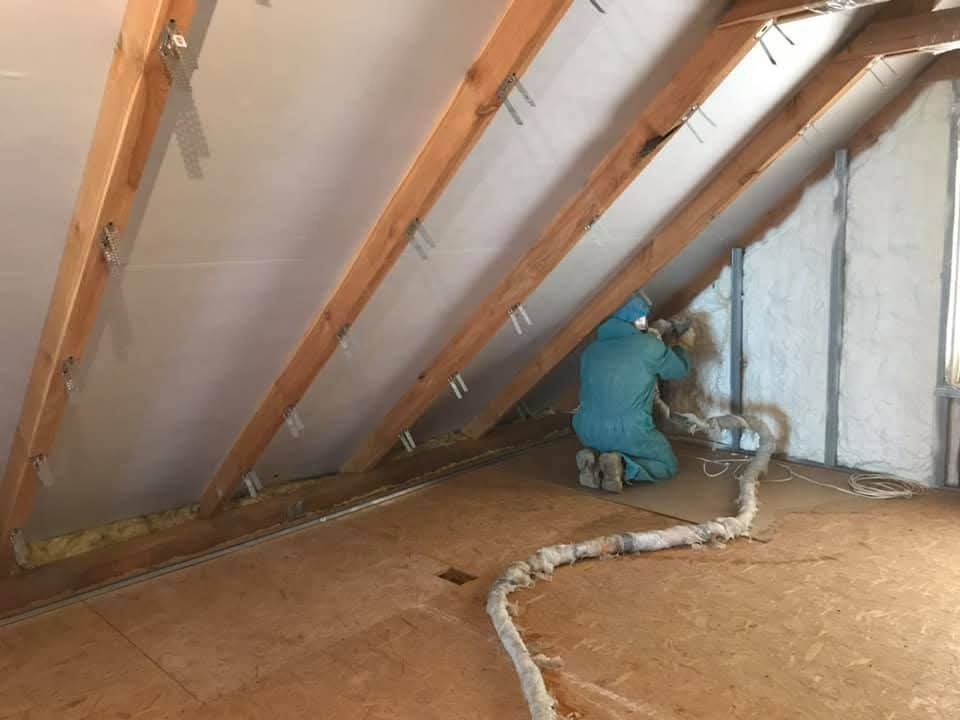 Pracownik w kombinezonie aplikuje pianę izolacyjną na poddaszu, widoczne drewniane krokwie i fragmenty ocieplonej ściany. Wąż doprowadzający pianę leży na podłodze.