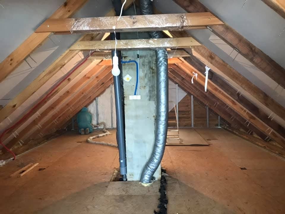 Poddasze z drewnianą konstrukcją, widoczny pracownik w kombinezonie podczas prac izolacyjnych. Elementy instalacji wentylacyjnej i oświetlenie.