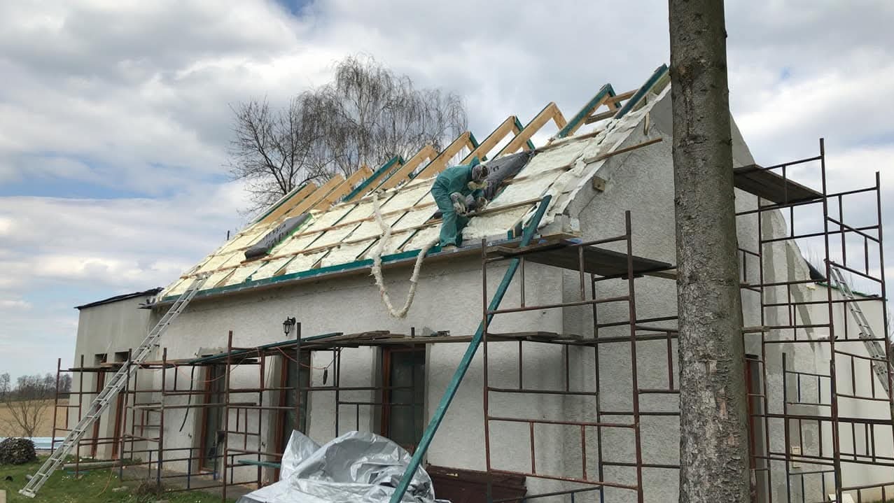 Pracownik w kombinezonie aplikuje pianę na dach domu jednorodzinnego ze rusztowania. Widoczne krokwie i częściowo ocieplona powierzchnia dachu. Praca w toku.