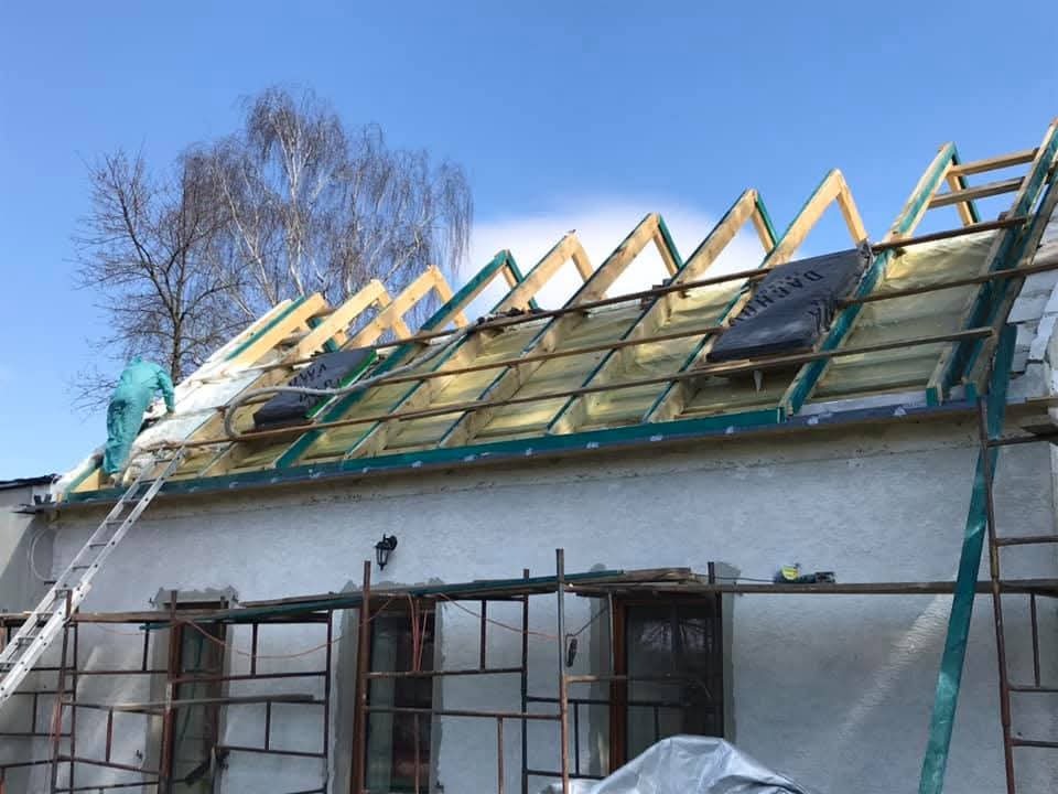Prace na dachu: montaż izolacji termicznej między krokwiami. Widoczny pracownik w kombinezonie ochronnym na drabinie. Dach pokryty folią i częściowo ocieplony.
