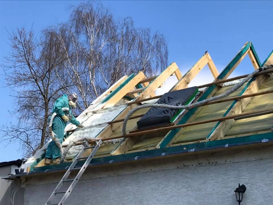 Pracownik w kombinezonie ociepla pianką PUR dach domu z drewnianą konstrukcją, stojąc na drabinie. Widoczne niebo i drzewo bez liści w tle.