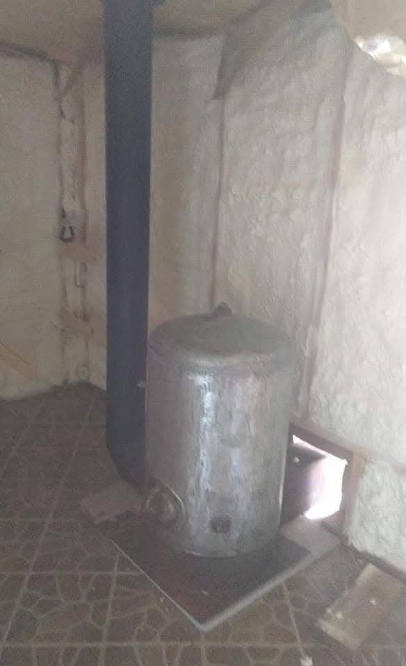 Wnętrze po ociepleniu pianką: ściany i sufit pokryte grubą warstwą izolacji, widoczny piec z czarnym kominem. Nieregularna faktura pianki, szara podłoga.