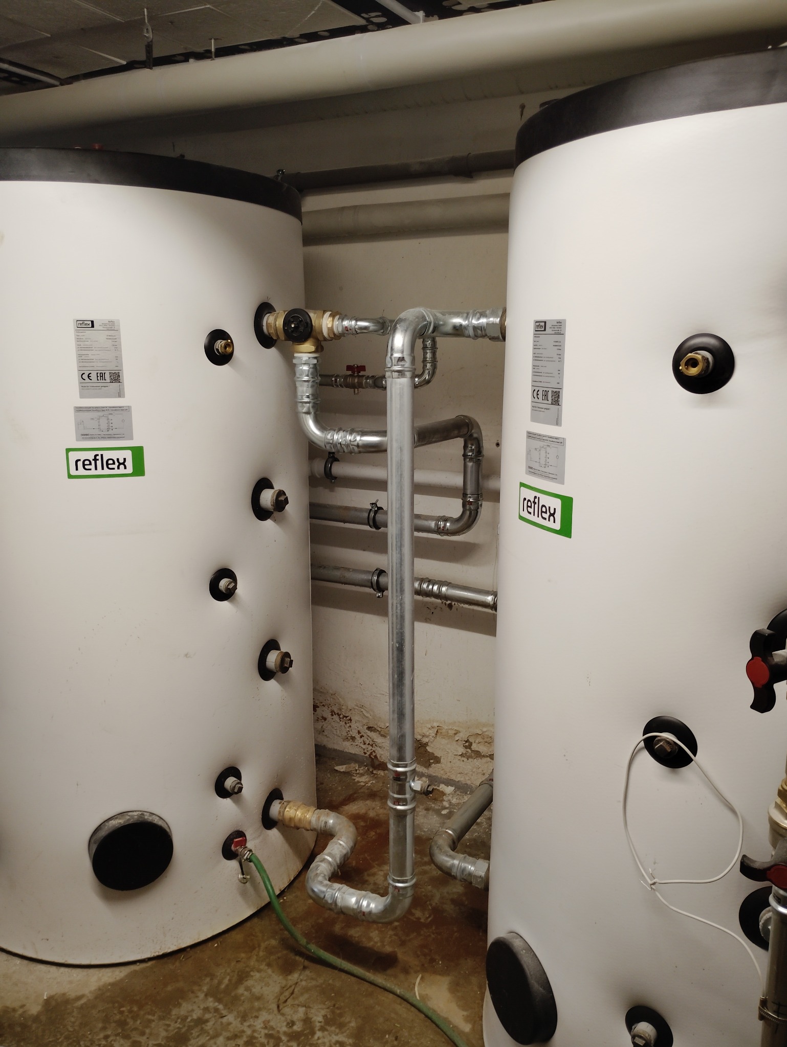 Dwa białe zbiorniki Reflex połączone rurami w kotłowni. Widoczne zawory, przyłącza i etykiety. Podłoga betonowa, ściany z widocznymi śladami wilgoci.