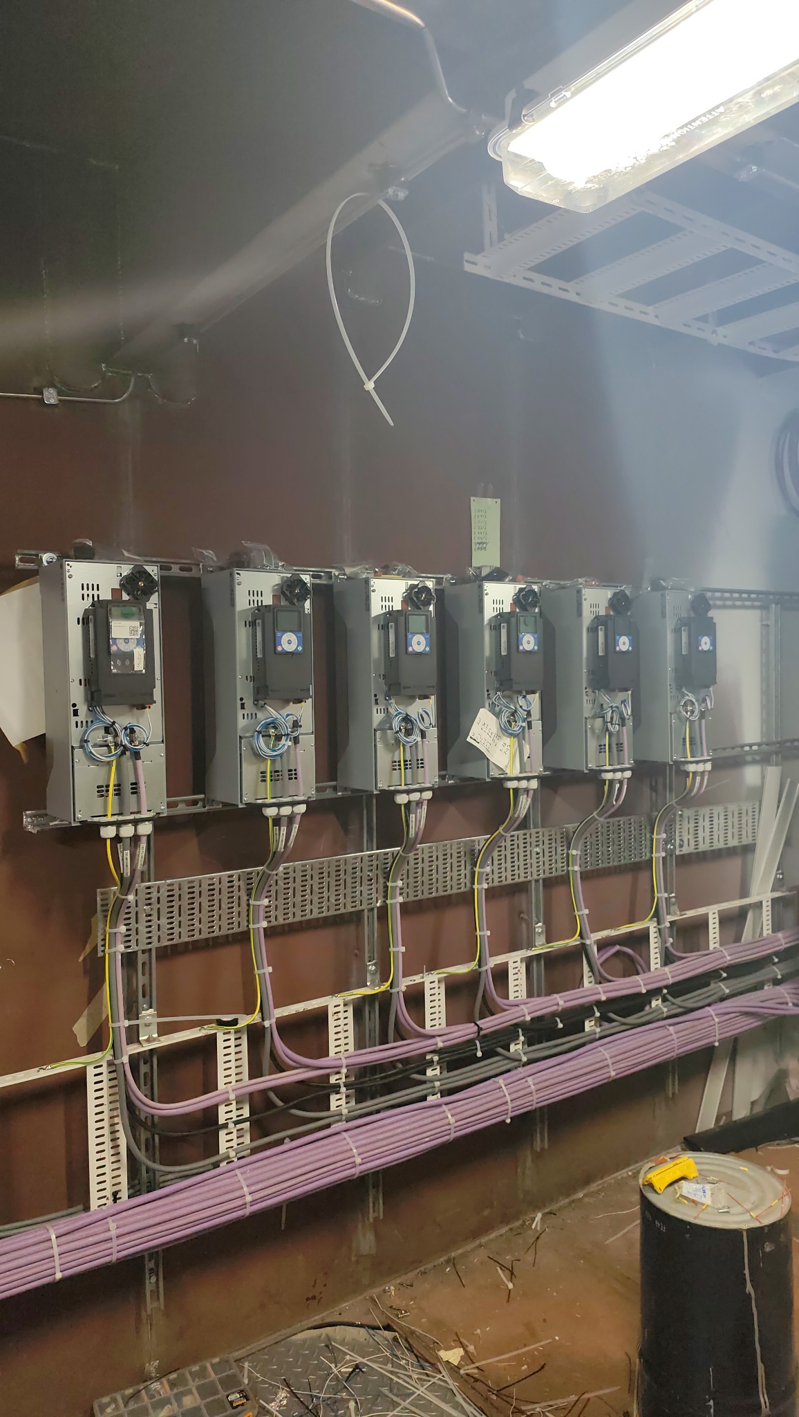 Rząd szarych szaf sterowniczych z podłączonymi kablami w kolorach żółtym i fioletowym, zamontowanych na metalowych szynach w pomieszczeniu technicznym.