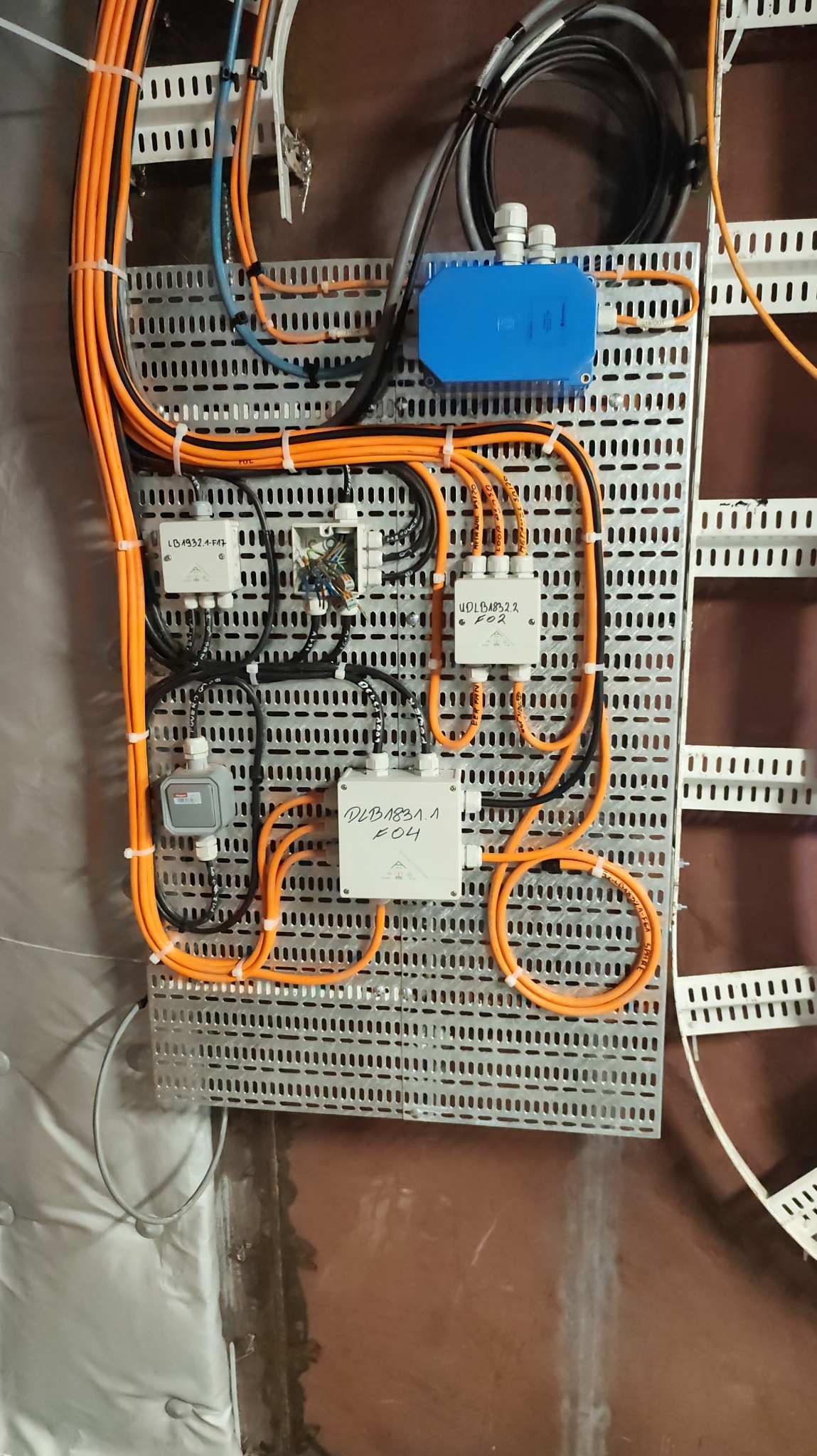 Szafa sterownicza z pomarańczowymi kablami, zamontowana na perforowanej płycie. Widoczne białe puszki z oznaczeniami i niebieska skrzynka. Instalacja w budynku.