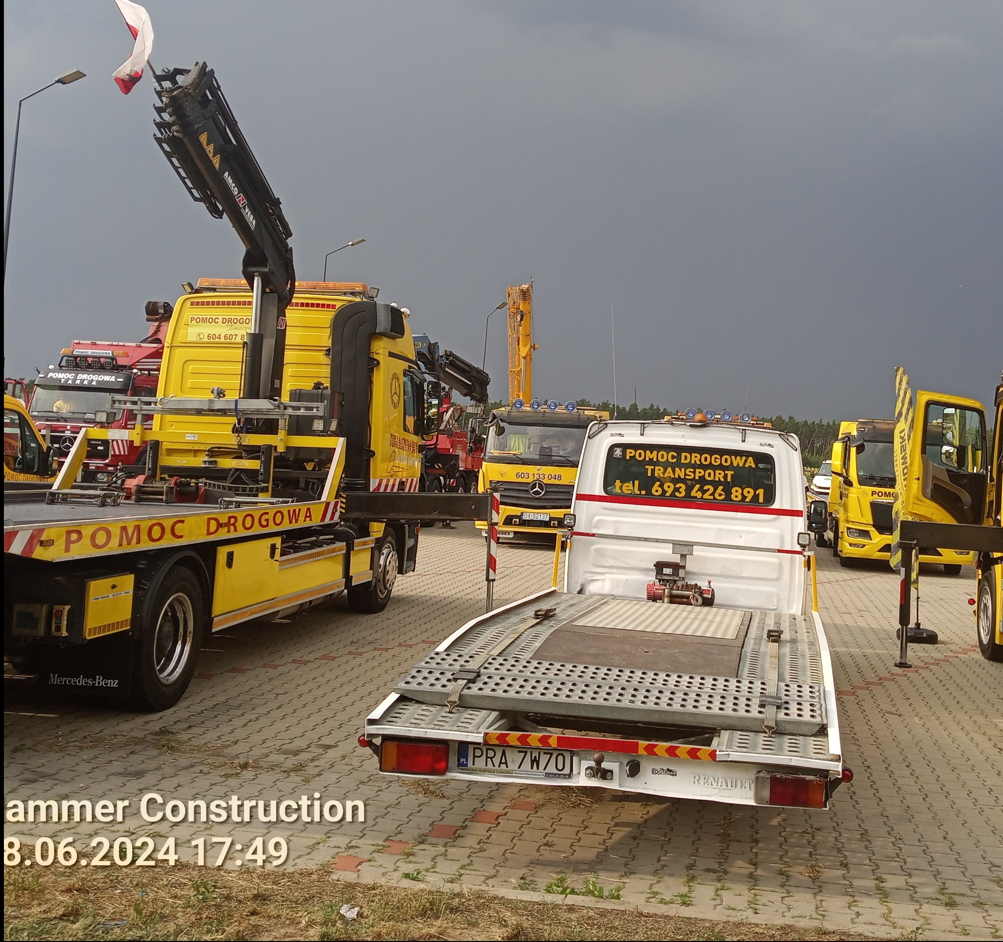 Flota pojazdów pomocy drogowej, w tym żółta laweta Mercedes-Benz z podniesionym ramieniem dźwigu i biała laweta Renault z platformą załadunkową, na tle pochmurnego nieba.