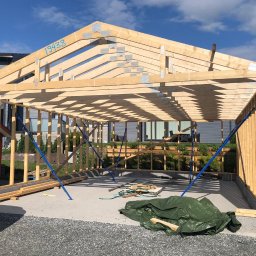 Mariusz - Konstrukcja szkieletowa w Lipno, widok z zewnątrz. Drewniane elementy połączone metalowymi płytkami, niebieskie podpory stabilizują konstrukcję. Widoczne fundamenty.