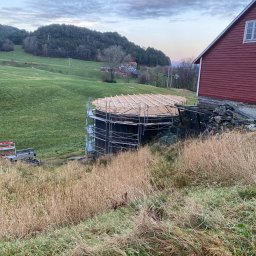 Mariusz - Budowa okrągłej konstrukcji z rusztowaniem na tle zielonego wzgórza i czerwonego budynku. Widoczne drewniane elementy konstrukcyjne.