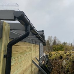 Mariusz - Nowe orynnowanie z czarnego PCV na drewnianej konstrukcji z szarymi elementami, widok z dołu pod kątem. Na dachu widoczna przezroczysta płyta. Pochmurne niebo w tle.