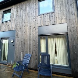 Mariusz - Nowoczesny dom szkieletowy z drewnianą elewacją i tarasem. Dwa niebieskie krzesła na tarasie. Okna z białymi zasłonami. Ciemna kolorystyka.