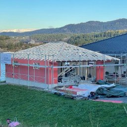 Mariusz - Budowa domu szkieletowego w malowniczej scenerii: konstrukcja z czerwonymi panelami, drewniany szkielet dachu, rusztowania i przyczepa na tle jeziora i gór.