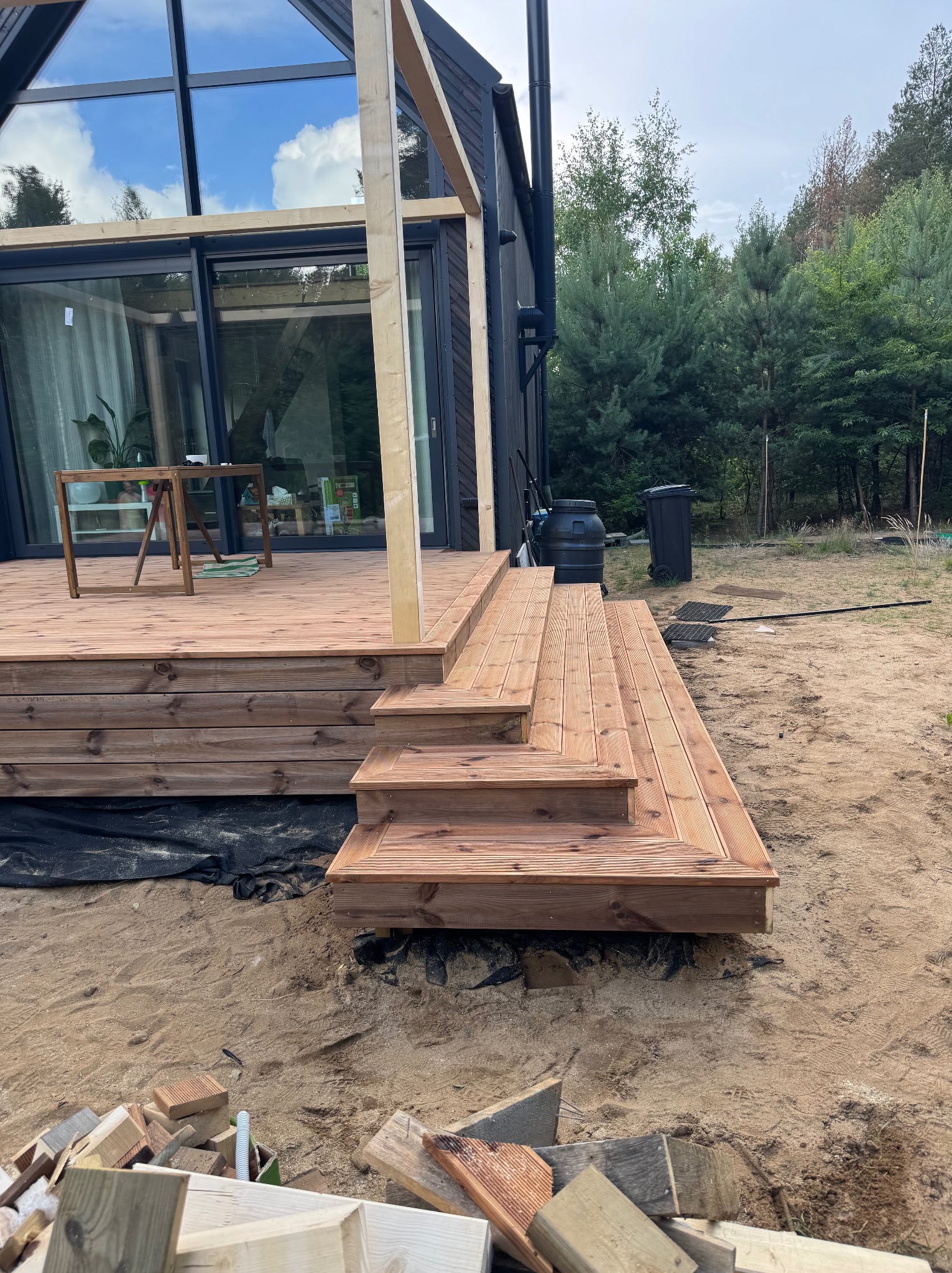 Nowoczesny taras z drewna z niskimi schodami, częściowo zadaszony, w trakcie budowy przy domu z dużymi oknami. Widoczne elementy konstrukcyjne i narzędzia.