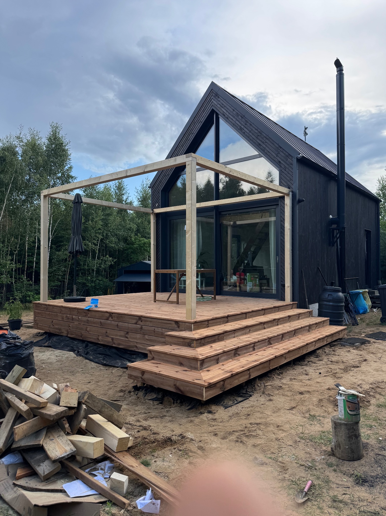 Nowoczesny dom z czarną elewacją i drewnianym tarasem w trakcie budowy. Widoczna konstrukcja pergoli i schody tarasowe. W tle las i pochmurne niebo.