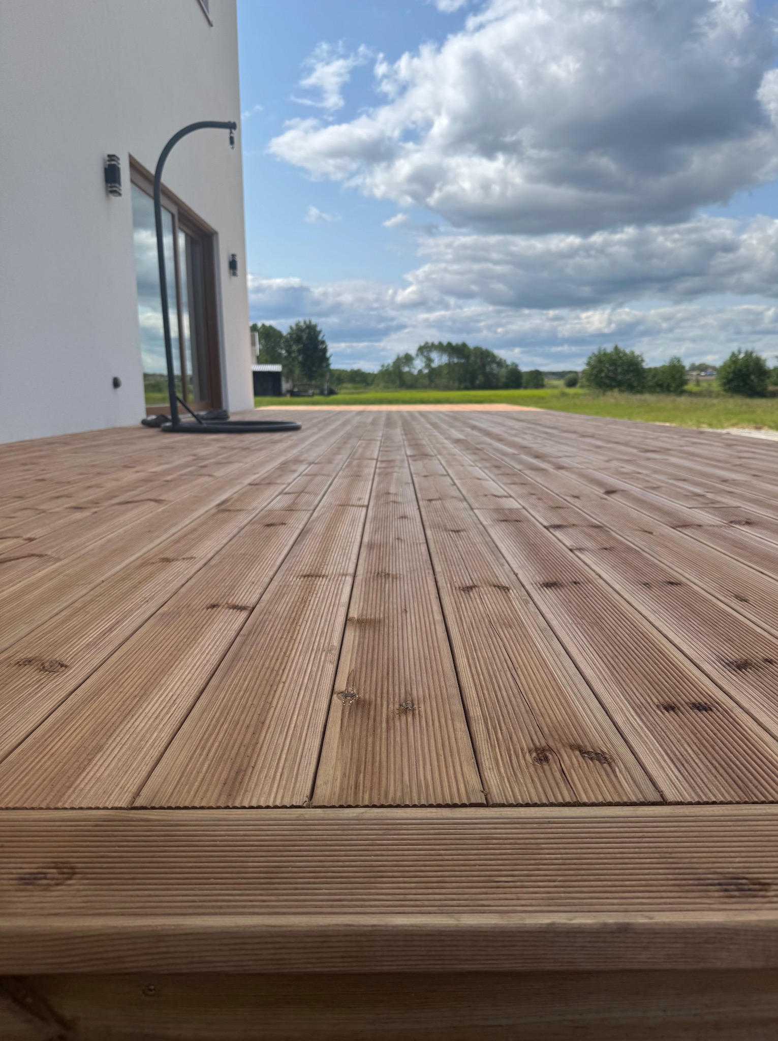 Szeroki, niski kadr na drewniany taras z ryflowanymi deskami, przylegający do białego budynku z widocznym fragmentem okna i elementem konstrukcyjnym. W tle zielony krajobraz.