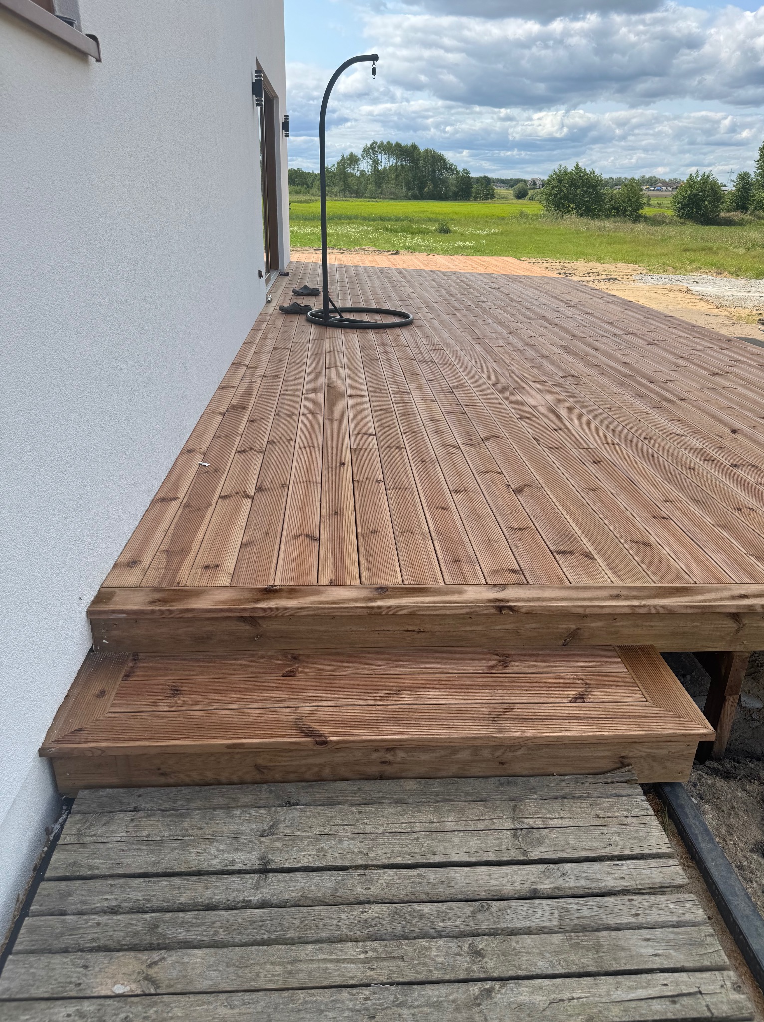 Nowy taras drewniany z dwoma stopniami przy białej ścianie domu, z widokiem na zielone pole i błękitne niebo. Widoczne deski tarasowe i konstrukcja schodów.