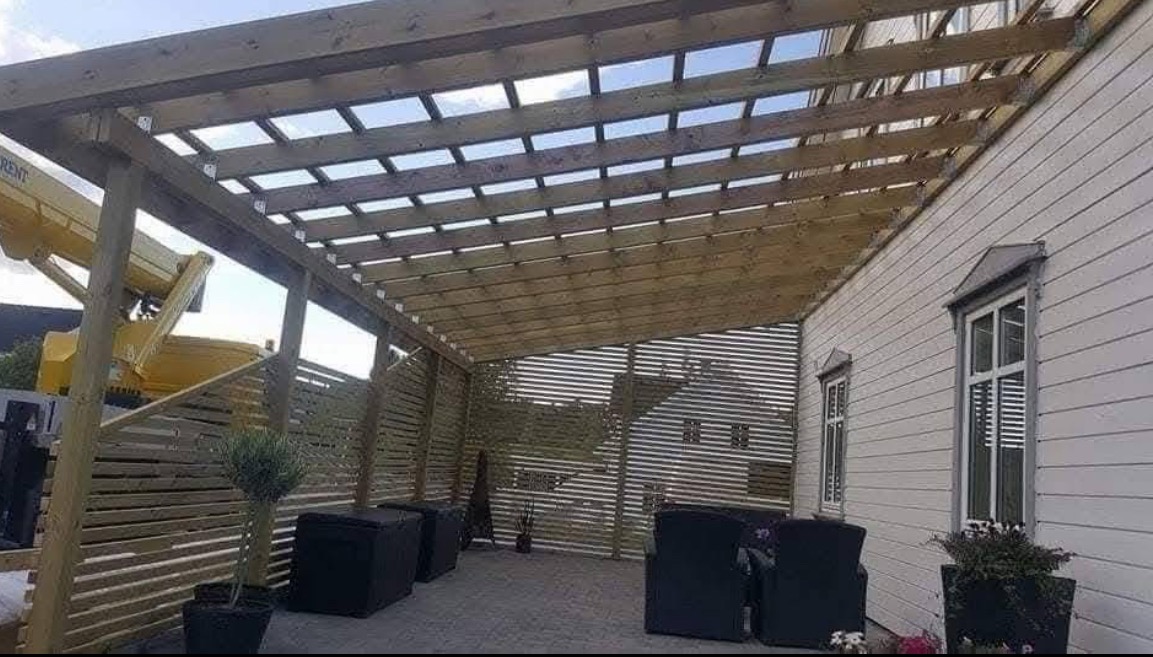 Drewniana pergola z ażurowym dachem i ścianami, przylegająca do budynku z białymi panelami. W tle widoczny podnośnik koszowy.