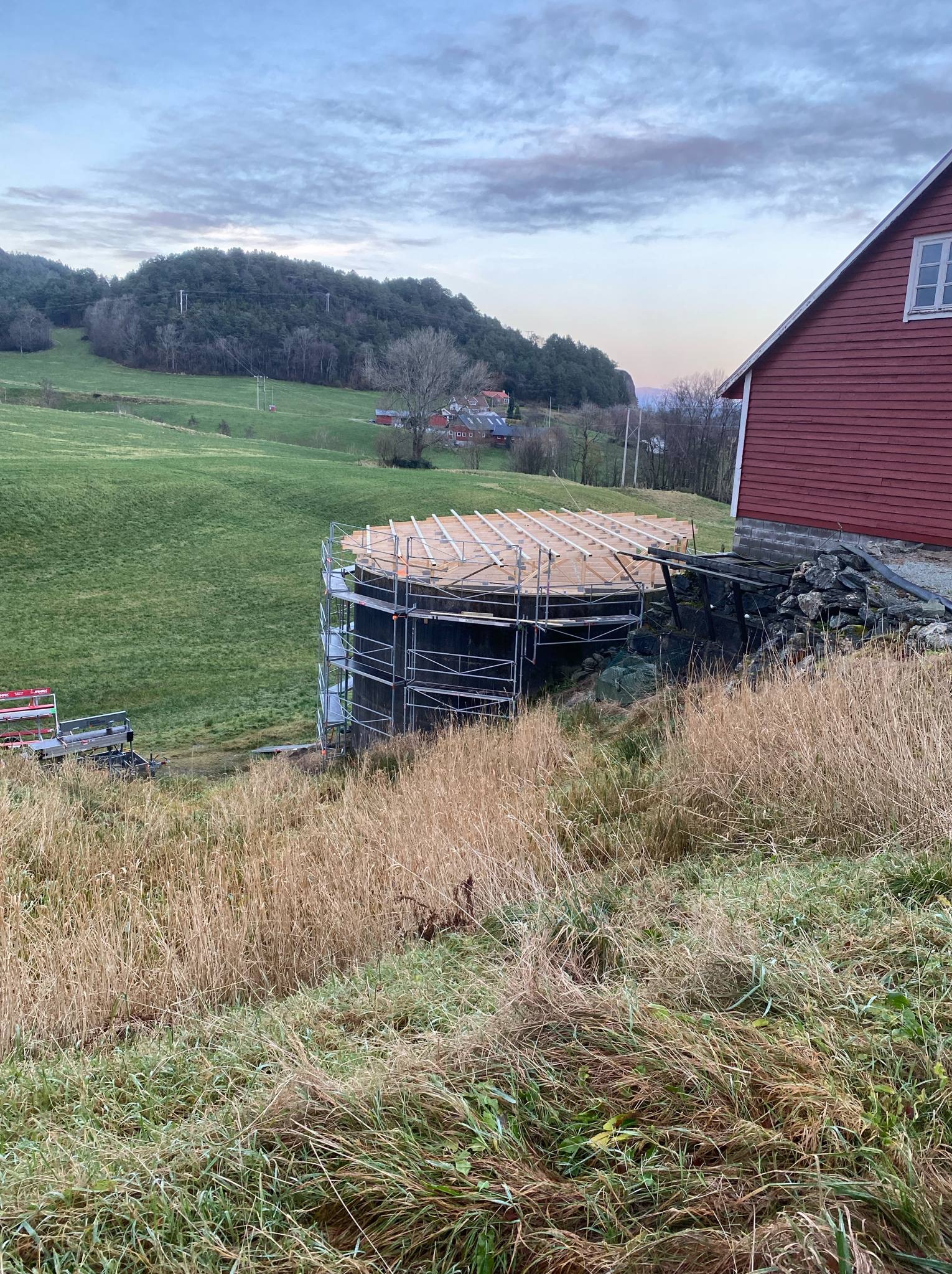 Budowa okrągłej konstrukcji z rusztowaniem na tle zielonego wzgórza i czerwonego budynku. Widoczne drewniane elementy konstrukcyjne.