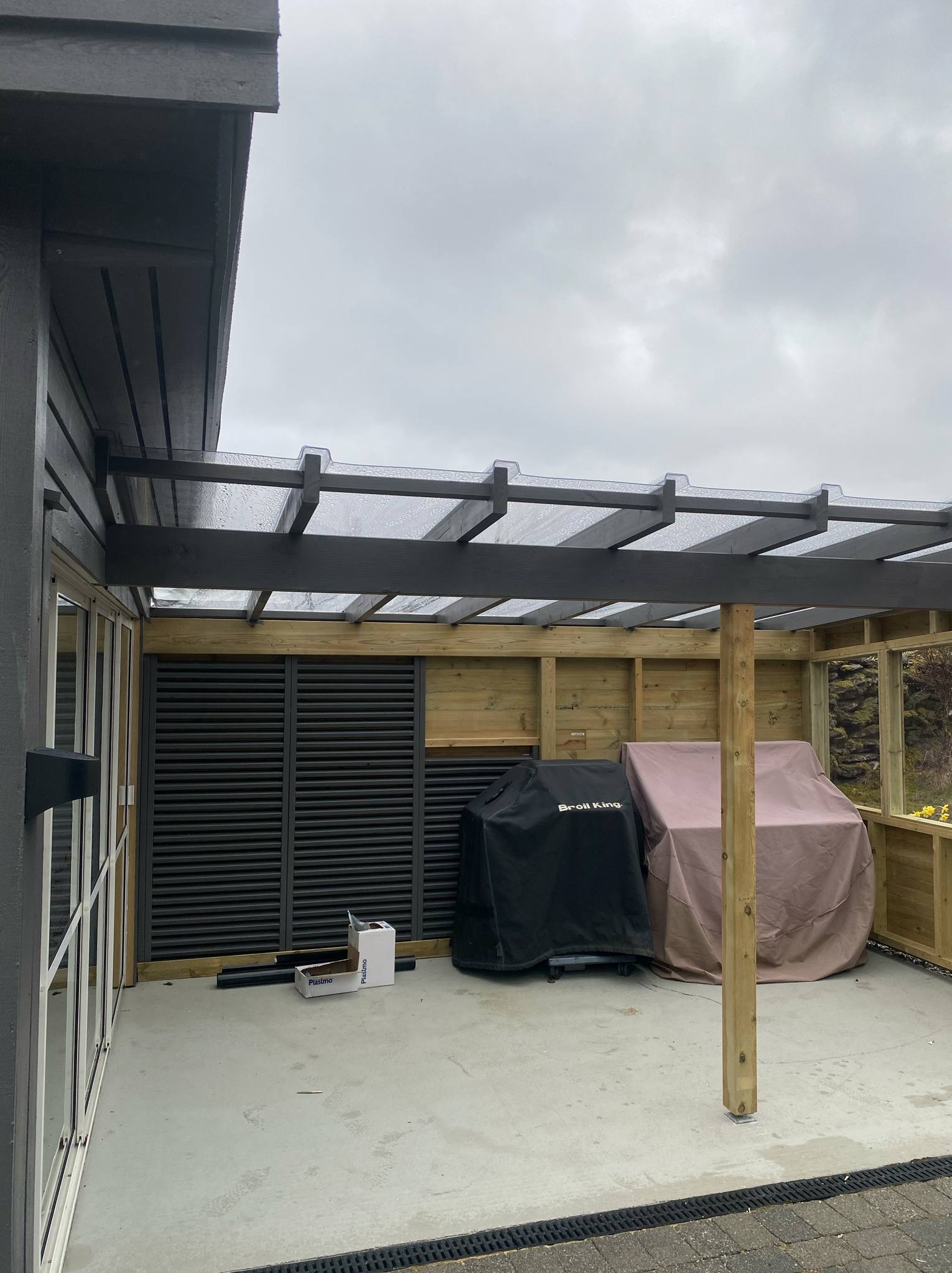 Zadaszony taras z drewnianą konstrukcją i szarymi żaluzjami. Dwa grille pod pokrowcami. Widoczne elementy konstrukcyjne i wykończeniowe. Betonowa podłoga z odpływem liniowym.