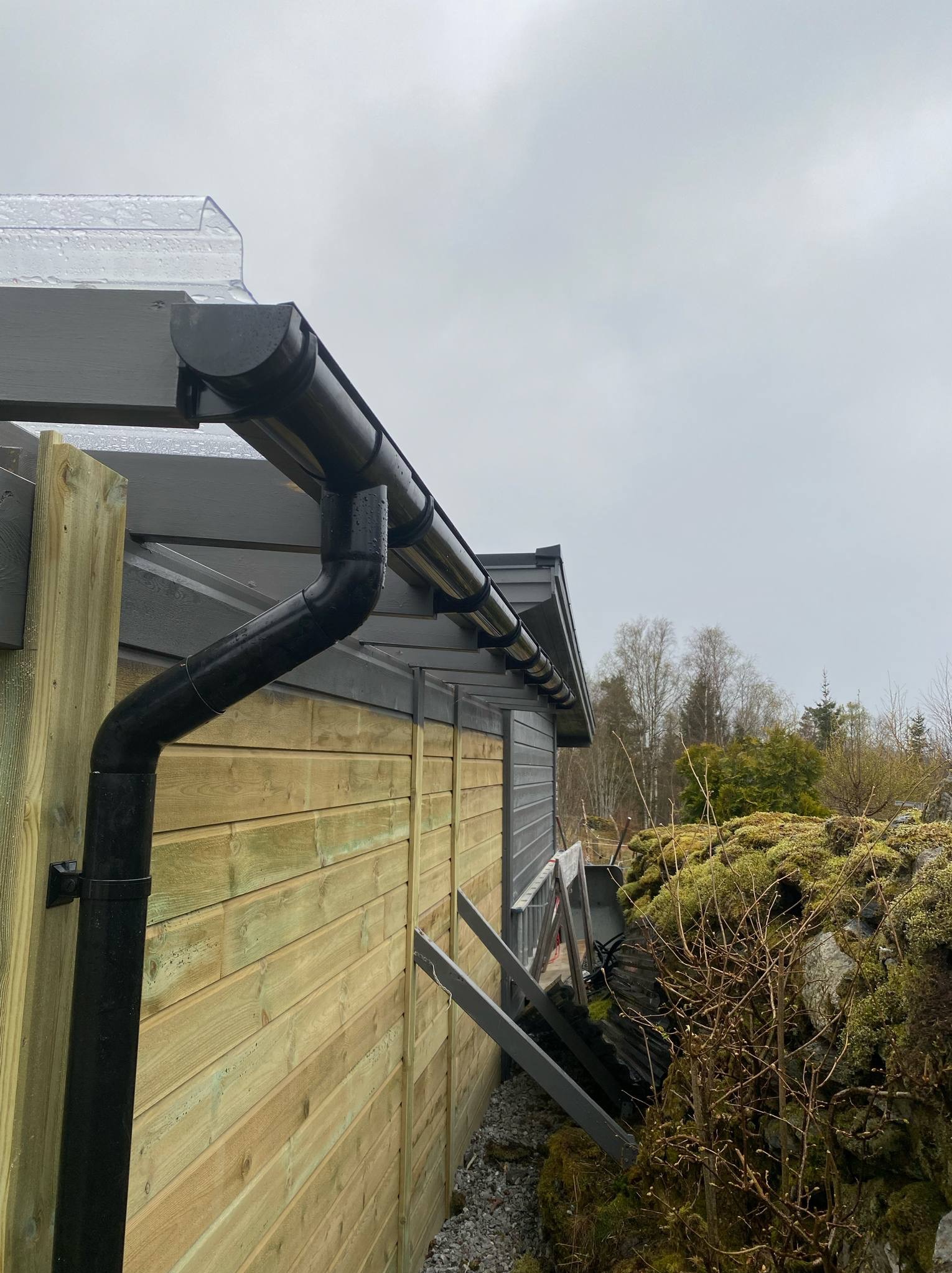 Nowe orynnowanie z czarnego PCV na drewnianej konstrukcji z szarymi elementami, widok z dołu pod kątem. Na dachu widoczna przezroczysta płyta. Pochmurne niebo w tle.