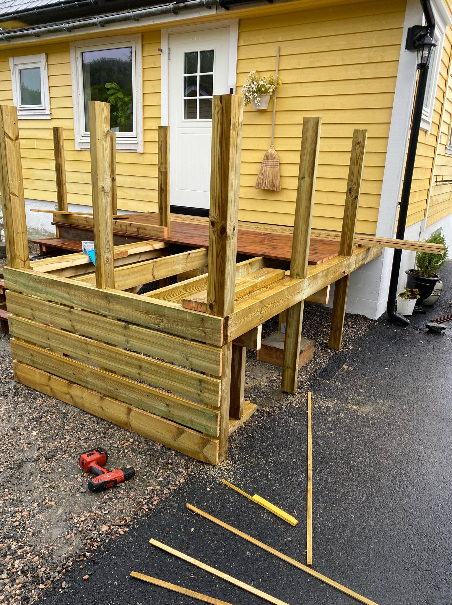Konstrukcja drewnianego tarasu w trakcie budowy przy żółtym domu. Widoczne narzędzia i elementy konstrukcyjne. Częściowo ułożona podłoga tarasu.